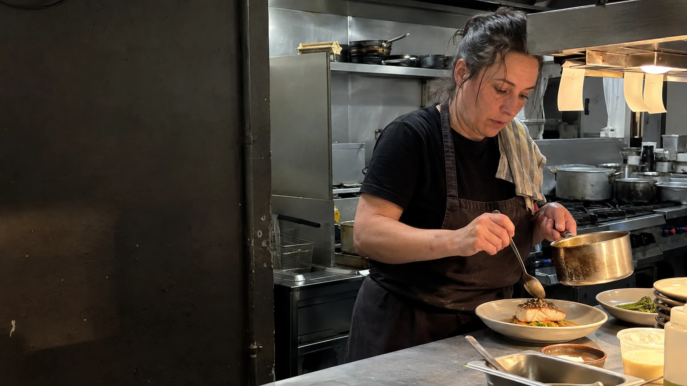 A chef-owner spooning sauce over a plated dish at the pass during service