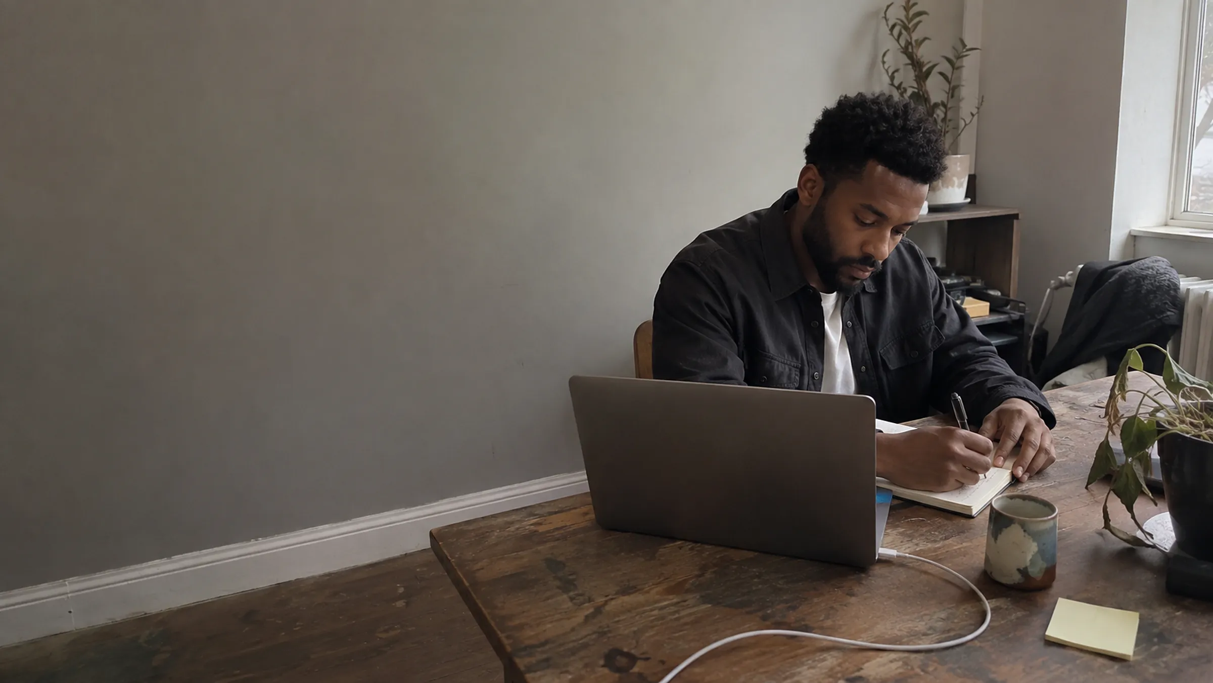 A young founder writing in a notebook at a worn wooden table in a small studio office